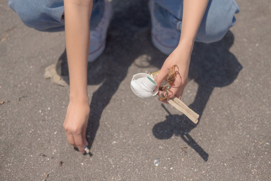 A person crouching on a paved outdoor surface, holding a small homemade device consisting of a white spherical object, possibly made of plastic or foam, attached to a wooden stick or handle. The person's hands are visible, with one hand gripping the device and the other hand resting on the ground. They are wearing light blue jeans and white sneakers, and the background features a textured asphalt surface with small stones and debris. The scene is lit by natural daylight, emphasizing the textures of the materials and the casual outdoor environment, which may relate to informal waste collection or clean-up activities managed by local waste services such as Rubbish Removal Earls Court.