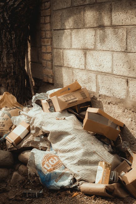 A pile of discarded cardboard boxes and used plastic packaging materials is stacked against a rough, weathered brick and concrete wall, situated outdoors on a dirt surface. The cardboard boxes vary in size, some are flattened while others still have folded edges, with printed labels and barcode markings visible on their surfaces. Among the debris, there is a white woven bag or sack partially filled or crumpled, and some of the packaging is torn or open, revealing only material waste. To the left side of the image, a portion of a tree trunk with dark, textured bark is visible, casting some shadows on the debris. The environment suggests a site for waste collection or disposal, potentially managed by a rubbish removal service like Rubbish Removal Earls Court, which handles independent waste clearance outside of municipal systems. The lighting appears natural, possibly from daylight, with a neutral tone emphasizing the utilitarian nature of the scene and the importance of proper waste management for private or commercial rubbish removal.