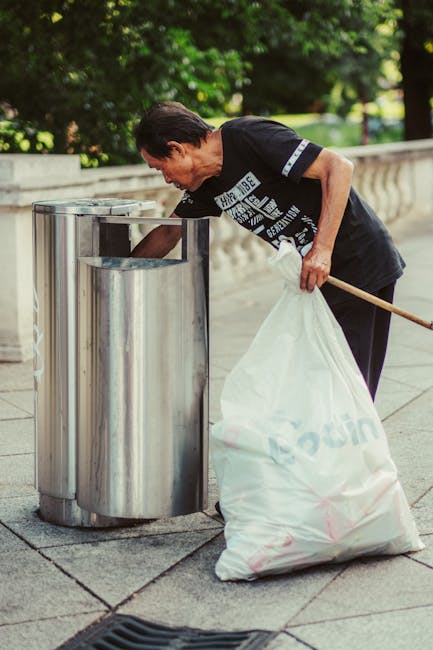 A woman with short dark hair, wearing a black T-shirt with a graphic print and dark trousers, is seen standing outdoors on a paved area surrounded by green trees and foliage. She is leaning forward, actively disposing of waste into a cylindrical metal rubbish bin with a large rectangular opening at the top. She is holding a large white plastic bag filled with rubbish in her left hand and using a wooden stick in her right hand to place waste into the bin. The scene suggests an informal or public rubbish collection, possibly as part of a community cleaning or maintenance activity. The waste appears to be general household or street refuse, and the metal bin has a clean, polished finish with a modern design. This scene highlights practices associated with rubbish removal and waste management in public spaces, with the individual performing an on-site waste disposal action which aligns with private or independent rubbish collection services offered by companies like Rubbish Removal Earls Court.