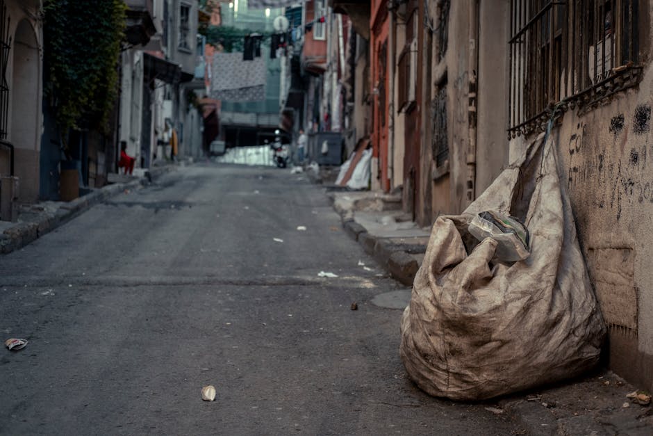 A large, worn and dirty white plastic bag filled with mixed rubbish, resting on a concrete pavement along a narrow urban street. The bag is loosely tied at the top, revealing some discarded packaging and materials spilling out. The street is lined with closely packed residential buildings featuring metal security bars on windows and graffiti or grime on the walls. In the background, laundry is hung on lines strung between buildings, and the environment appears somewhat neglected with scattered litter and debris, including small paper pieces and cans. The lighting suggests an overcast day or shaded area, and the scene reflects an instance of informal rubbish accumulation, which could be indicative of limited access to regular waste collection services. The image illustrates a typical scenario where private waste handling, such as bagging rubbish for potential collection, is occurring in a densely built area, aligning with the scope of rubbish removal services like those offered by Rubbish Removal Earls Court.