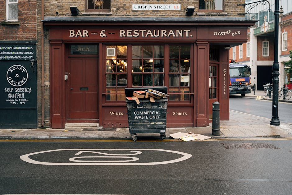 A black wheeled commercial waste bin with bold white lettering that reads 'Commercial Waste Only' is positioned on the sidewalk in front of a red brick storefront with wooden framing, large glass windows, and a sign advertising a bar and restaurant. The bin is overflowing with flattened cardboard boxes and paper, with some debris spilling onto the pavement. To the right of the bin, a small black bollard separates the sidewalk from the street, where a blue delivery truck is parked nearby, with bicycles secured to a rack further down. The storefront has a brown awning with white text indicating various services, and adjacent to it, a blackboard sign advertises a self-service buffet. The scene captures an urban setting during daylight, with a street sign reading 'Crispin Street' above the shop entrance, and a road marking visible in the foreground, indicating a parking or traffic regulation area. This image illustrates the context of private waste collection and rubbish disposal services often used by local businesses in the area.