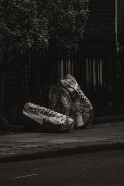 In the foreground, three large black plastic rubbish bags are visibly placed on a paved sidewalk next to a curb, with the bags appearing full and slightly crinkled, indicating they contain waste or discarded items. The bags are positioned adjacent to a black metal fence with vertical bars, and behind the fence, there is foliage from trees or bushes, providing a natural background contrast. The scene is outdoors, likely in an urban or residential area, with soft natural lighting suggesting an overcast day or shaded environment. The bags and the surrounding environment evoke a setting associated with private waste collection or rubbish disposal services, such as those offered by Rubbish Removal Earls Court, emphasizing the need for professional rubbish removal from commercial or residential premises. The overall mood is neutral and factual, with no additional objects or actions visible, aligning with an observational description suitable for accessibility purposes and SEO relevance in waste management content.