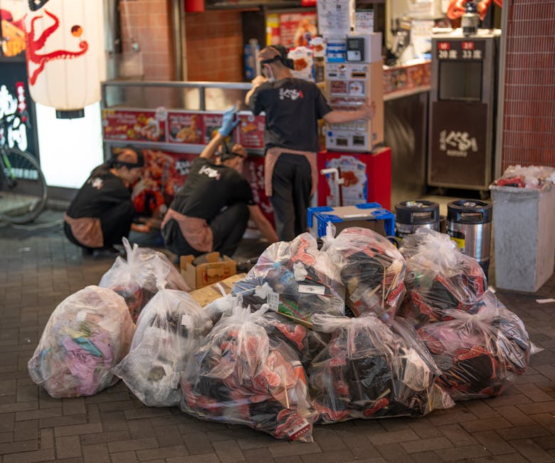 A black wheeled commercial waste bin with bold white lettering that reads 'Commercial Waste Only' is positioned on the sidewalk in front of a red brick storefront with wooden framing, large glass windows, and a sign advertising a bar and restaurant. The bin is overflowing with flattened cardboard boxes and paper, with some debris spilling onto the pavement. To the right of the bin, a small black bollard separates the sidewalk from the street, where a blue delivery truck is parked nearby, with bicycles secured to a rack further down. The storefront has a brown awning with white text indicating various services, and adjacent to it, a blackboard sign advertises a self-service buffet. The scene captures an urban setting during daylight, with a street sign reading 'Crispin Street' above the shop entrance, and a road marking visible in the foreground, indicating a parking or traffic regulation area. This image illustrates the context of private waste collection and rubbish disposal services often used by local businesses in the area.