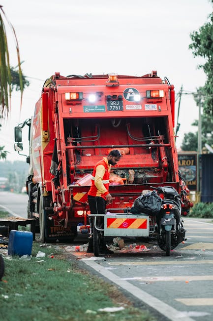 A worker wearing a red and yellow high-visibility vest is standing beside a large red rubbish collection truck on a street, operating the rear compactor mechanism. The truck's body is made of painted metal with visible bolts and hydraulic components, and the back features safety lights and reflective stripes. In front of the worker, there is a small metallic container attached to a black motorcycle parked close by, which has a black plastic bag placed on its seat. The scene is set outdoors during the daytime, with a partly cloudy sky and some greenery, including trees and grass along the roadside. The pavement is marked with white lines, indicating a designated area for waste collection or recycling. Behind the scene, there are street signs, utility poles, and a building with a sign partially visible, suggesting a commercial or mixed-use area. This image visually illustrates independent waste collection or private rubbish removal services, which are often used as alternative disposal methods to local authority services, supported by professional operators like Rubbish Removal Earls Court.