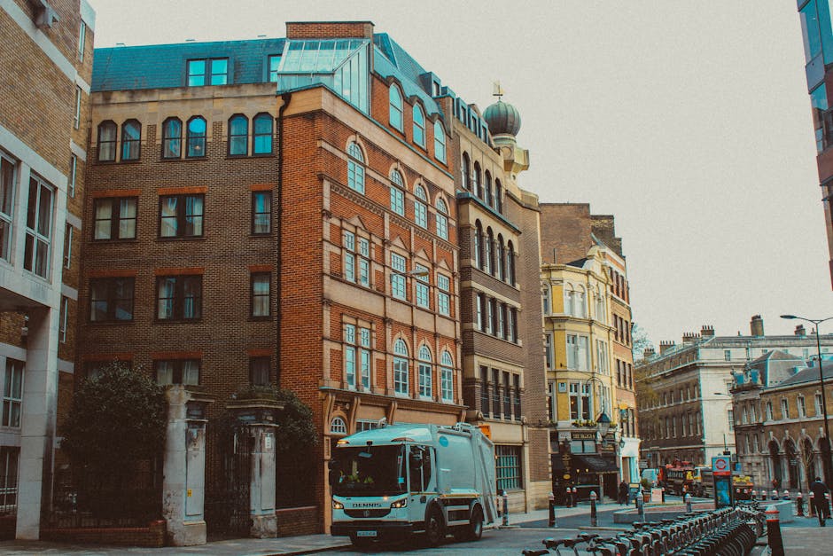 A street scene showing a grey rubbish collection van parked in front of a row of historic multi-storey buildings with mixed brick and stone facades. The buildings feature a variety of window styles, including arched and rectangular sash windows, with some having decorative stone detailing around the frames. The rooftops display different finishes, including slate and metal, with one building having a small dome-shaped turret topped with a cross. In the foreground, a low stone wall and iron gates mark an entrance to a building or courtyard, partially obscured by a small shrub. The street is lined with black bollards and bicycles are parked along a railing on the right side, while a few pedestrians and vehicles are visible in the distance, under an overcast sky. The scene subtly relates to private waste collection and rubbish removal services in an urban environment, fitting the context of independent waste management support for historic or commercial properties.