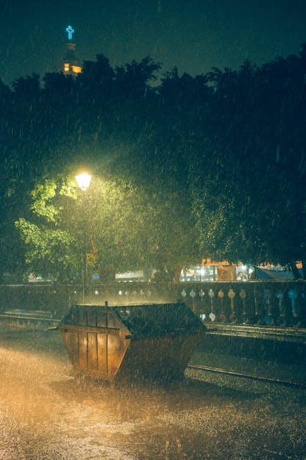 In the foreground of the image, there is a large, open-top dumpster made of rusted metal, positioned on a paved surface and overflowing with various waste materials, suggesting recent waste collection or ongoing rubbish disposal. Surrounding the dumpster, visible rainwater creates a reflective sheen on the ground, with raindrops visibly falling and splashing upon the pavement and the waste. Behind the dumpster, there is a row of dark green trees with dense foliage, which are illuminated by a nearby streetlamp emitting a warm, yellowish glow, partially diffusing through the rain and leaves. To the left, a traditional-style lamppost with a glowing light is present, providing illumination in the rainy, nighttime environment. In the background, a building with a cross on its top is faintly visible against the night sky, indicating a church or religious structure, with additional structures and lights further in the distance, under the overcast sky. The overall scene reflects a quiet urban setting during a heavy rainstorm, where private or independent waste disposal activities may be inferred without explicit action. Rubbish Removal Earls Court could be associated with such on-site waste handling scenarios in this context, emphasizing the importance of effective rubbish management during adverse weather conditions.